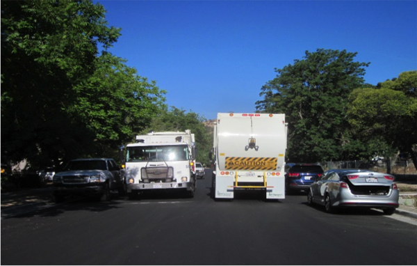 Two Paso Robles Waste Trucks travelling side-by-side, with cars parked on both sides of the street.